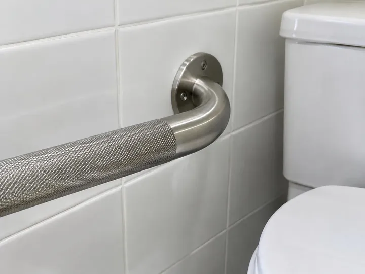 Knurled stainless steel grab bar with a round mounting flange on a tile wall beside a toilet