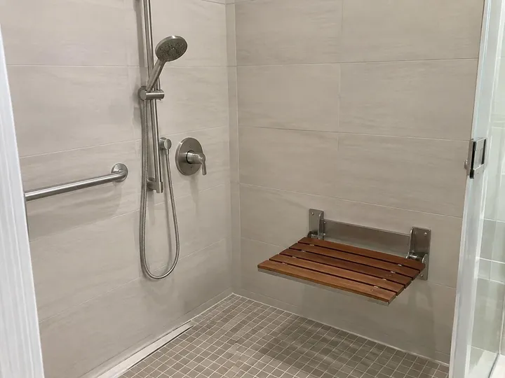 Wall-mounted fold-down teak shower seat beside a grab bar and handheld shower