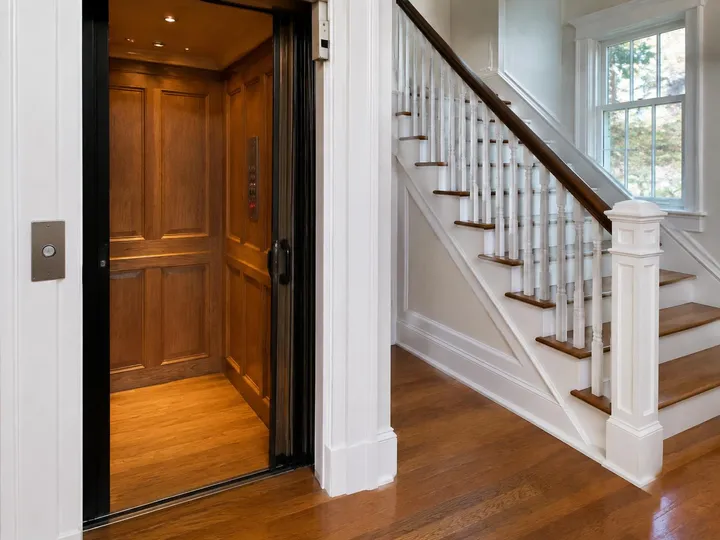 Home elevator with an open gate and wood-paneled cab next to a traditional staircase