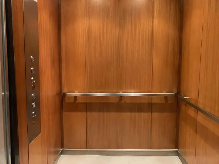 Inside a residential elevator cab with wood panels, a steel handrail, and call buttons
