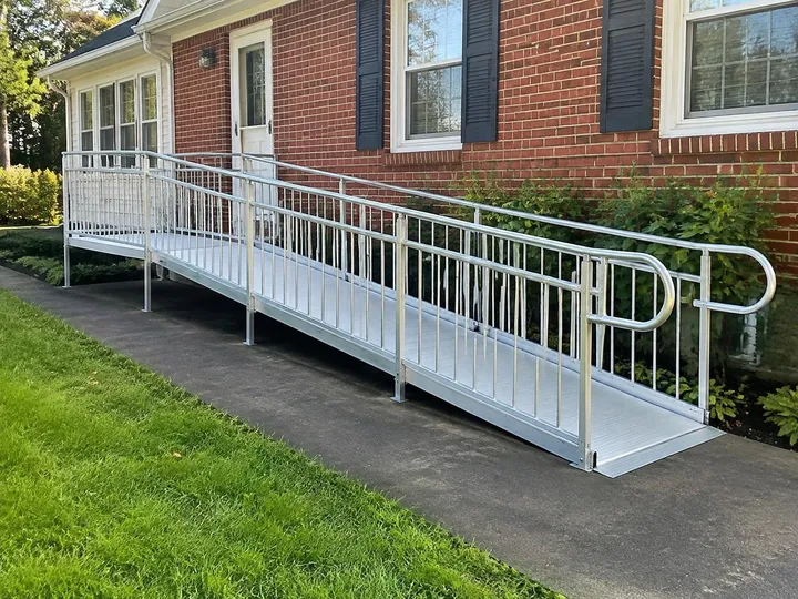 Long, gently sloped aluminum wheelchair ramp with handrails on both sides beside a brick house